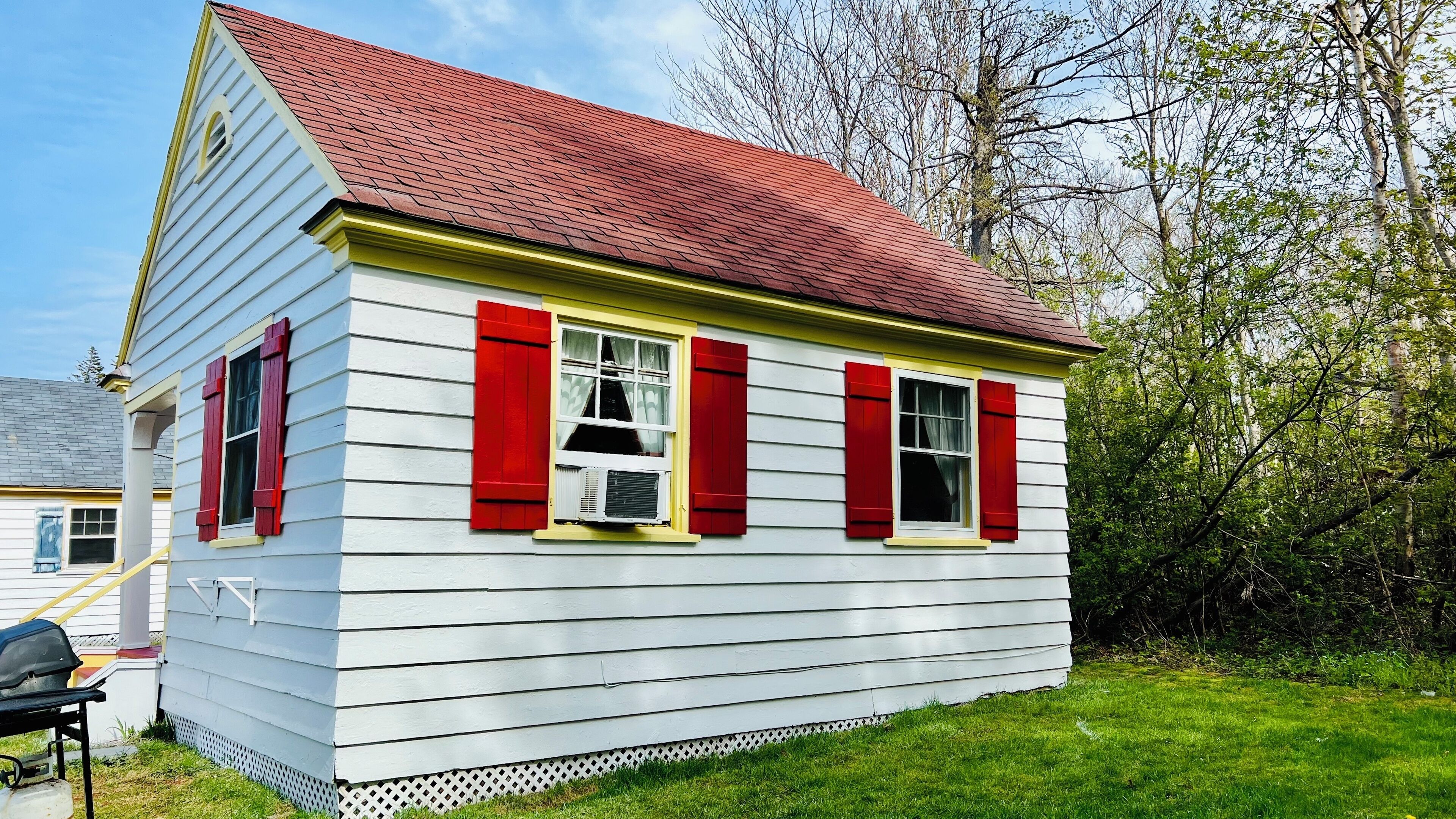 A small, single-story house with a red roof and white siding, featuring two windows with red shutters and a visible air conditioning unit.