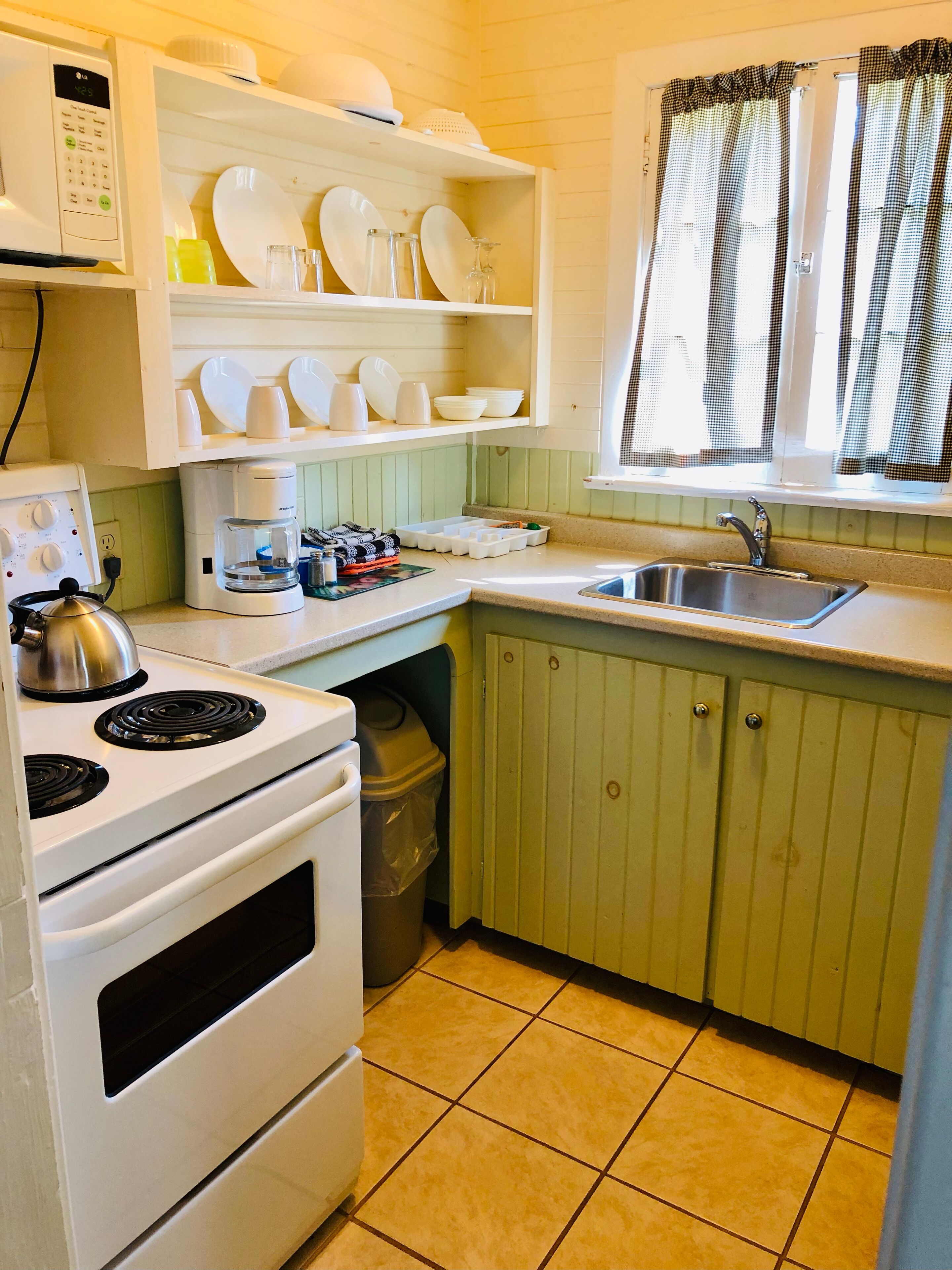 A compact kitchen with a stove, microwave, and sink. There are shelves above the counter holding dishes and glasses.