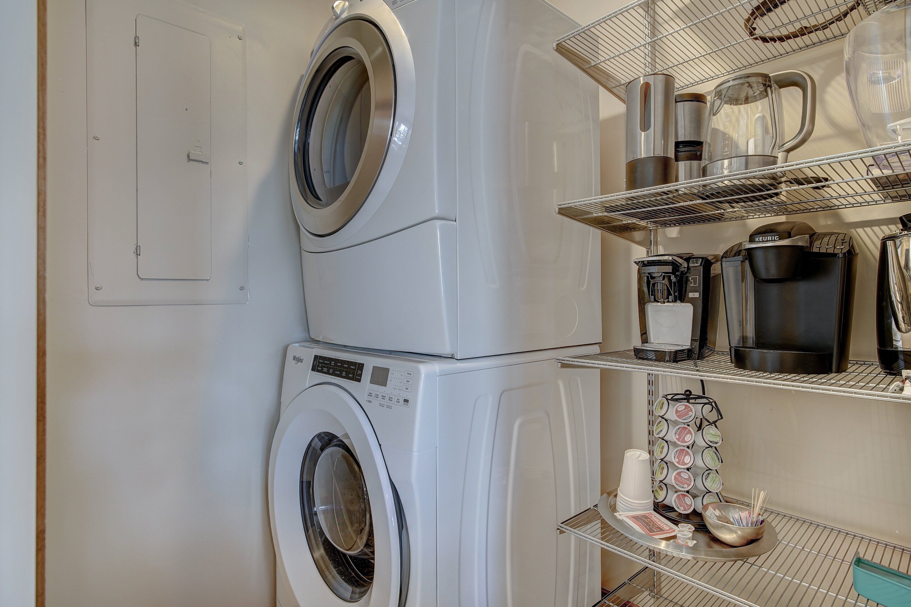 A laundry area with stacked washing machines and a shelf with coffee makers and cups.