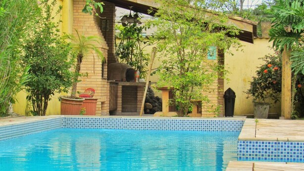 A swimming pool with a tiled edge, surrounded by greenery and a brick wall. - Hostel Brazil Backpackers (Guarujá)