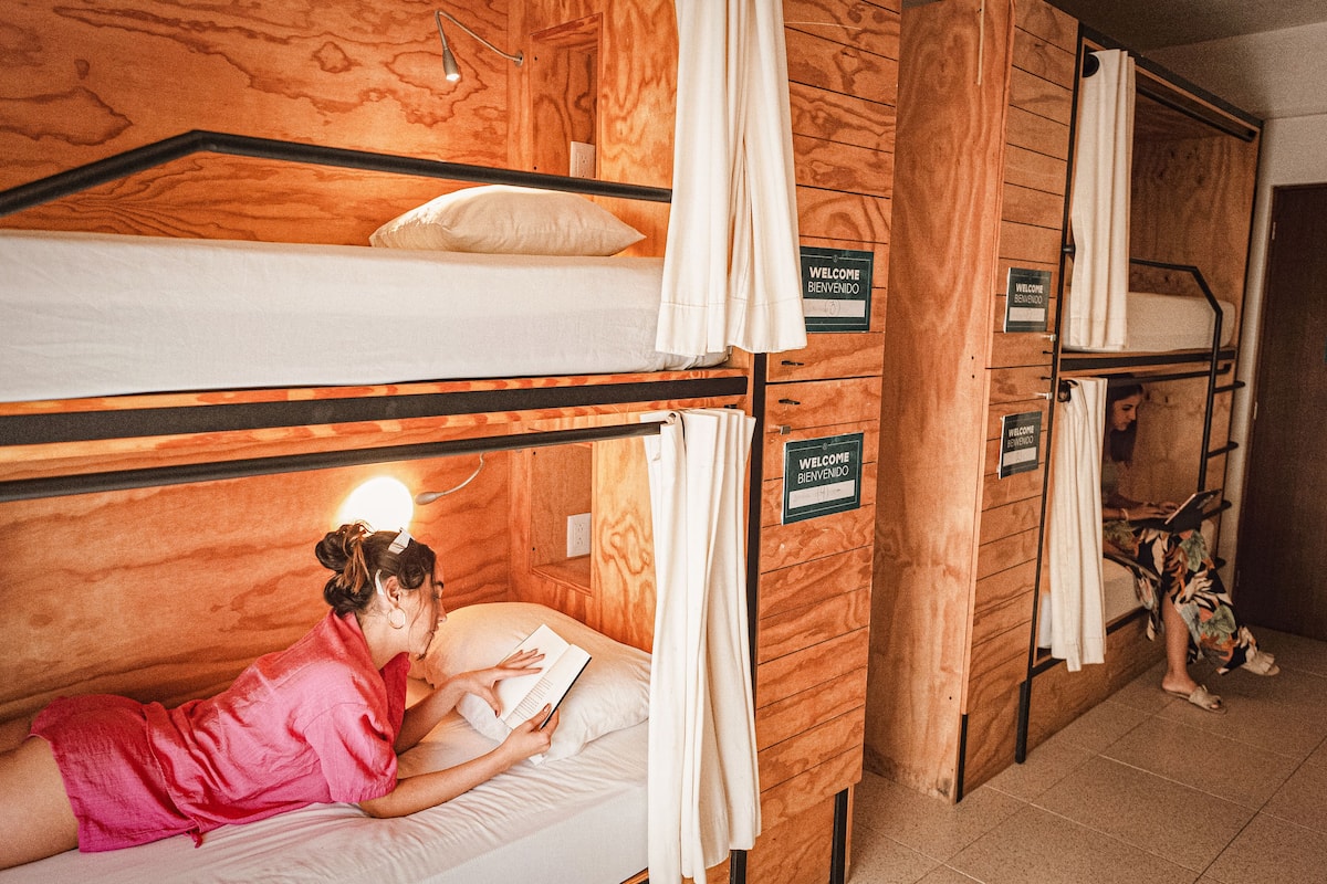 Une chambre avec des lits superposés, des murs en bois, un panneau d’accueil et une personne qui lit un livre.