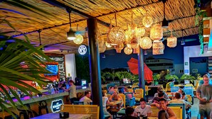 An indoor dining area with wooden tables and chairs, hanging light fixtures, and a sign indicating a beer brand. - Nomads Hotel & Rooftop pool (Cancun)