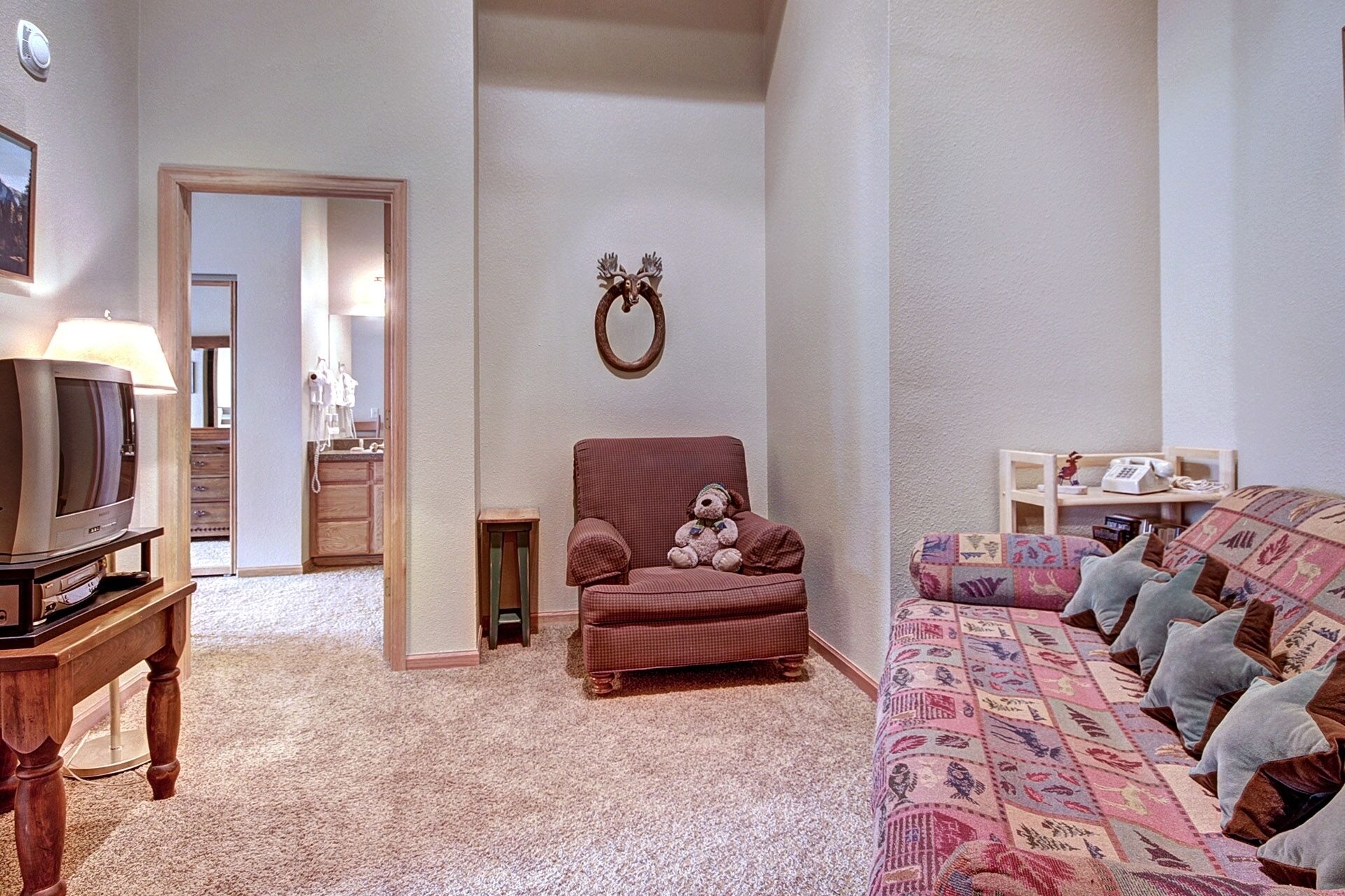 Una sala de estar con televisión, un sillón rojo, un sofá estampado y una mesita de madera.