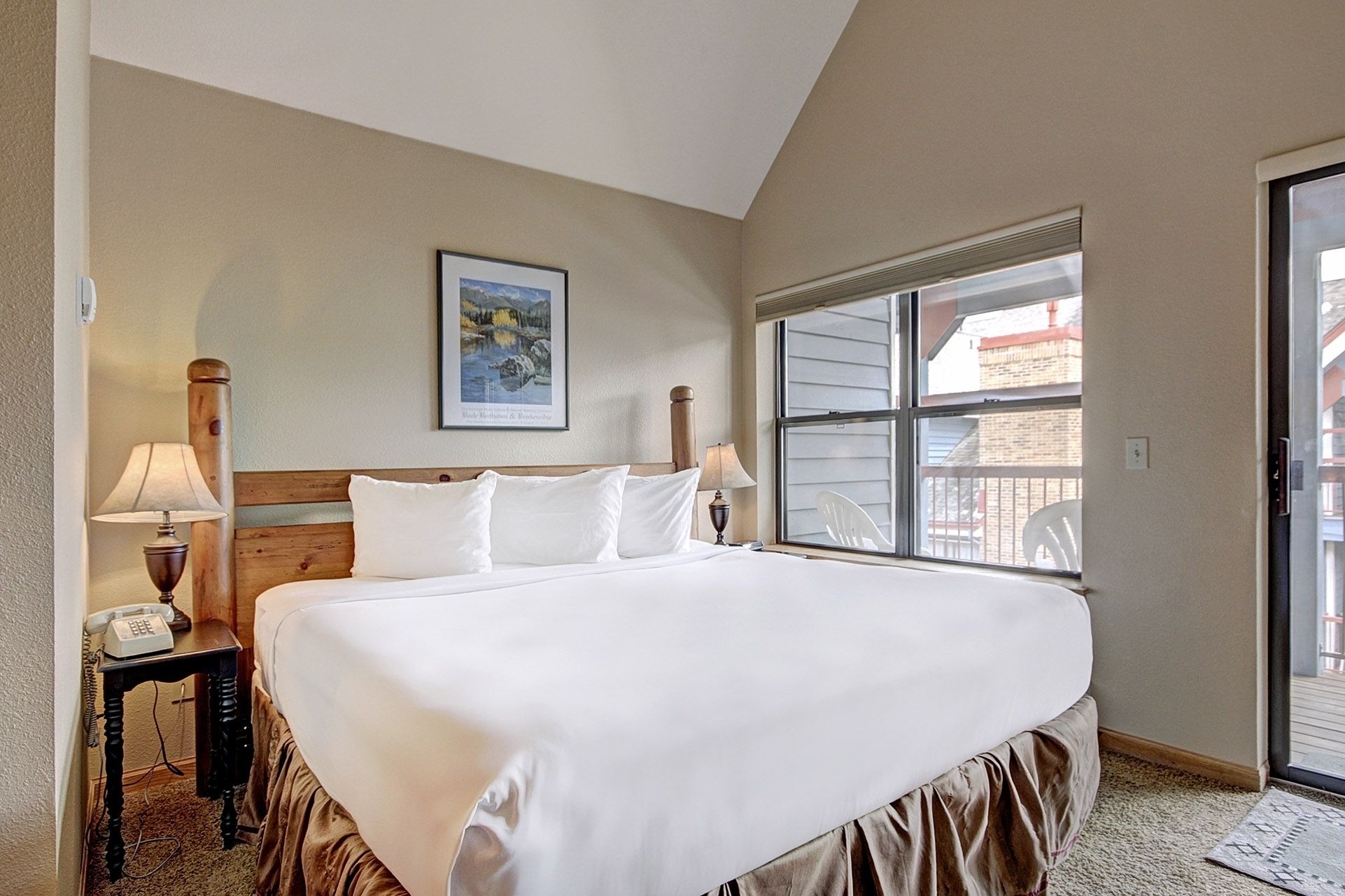 A hotel room with a large bed, a wooden headboard, a bedside lamp, a telephone, a window with a view of the outside, and a framed picture on the wall.