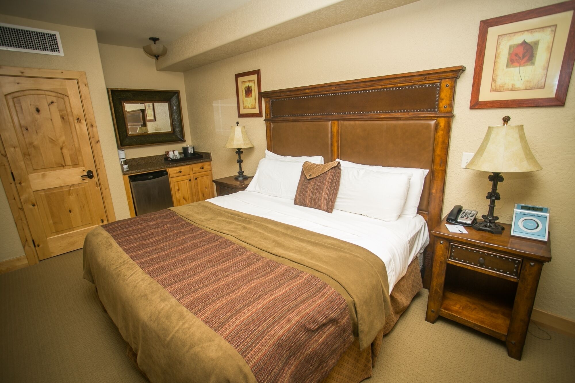 A hotel room with a large bed, wooden headboard, two bedside tables with lamps, a small desk, a telephone, and a framed picture on the wall.
