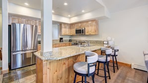 A modern kitchen with a stainless steel refrigerator, wooden cabinets, a granite countertop, and bar stools.