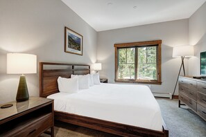 Un dormitorio ordenado con una cama grande, cabecera de madera, dos mesitas de noche con lámparas, un aparador y una ventana con vistas a zonas verdes.