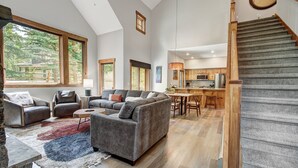 A spacious living area with a gray sectional sofa, a stone coffee table, and a large window overlooking a deck. A wooden staircase leads to a kitchen with modern appliances.