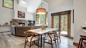 A spacious living area with a fireplace, a dining table with chairs, and large windows offering a view of the outdoors.