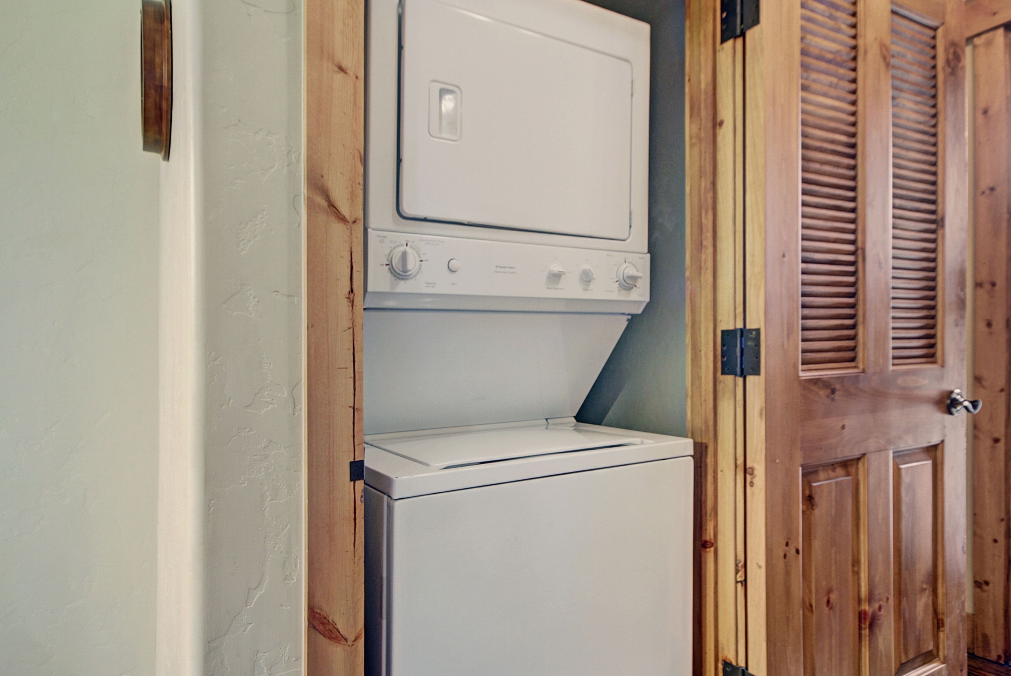 Une petite buanderie avec une machine à laver et un sèche-linge superposés, une porte en bois à panneaux ajourés et un mur blanc.