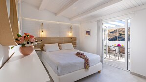 A modern bedroom with a large bed, a bedside table with a vase of flowers, a white headboard, and a balcony with a table and chairs. - Castello Apartments (Mylopotamos)