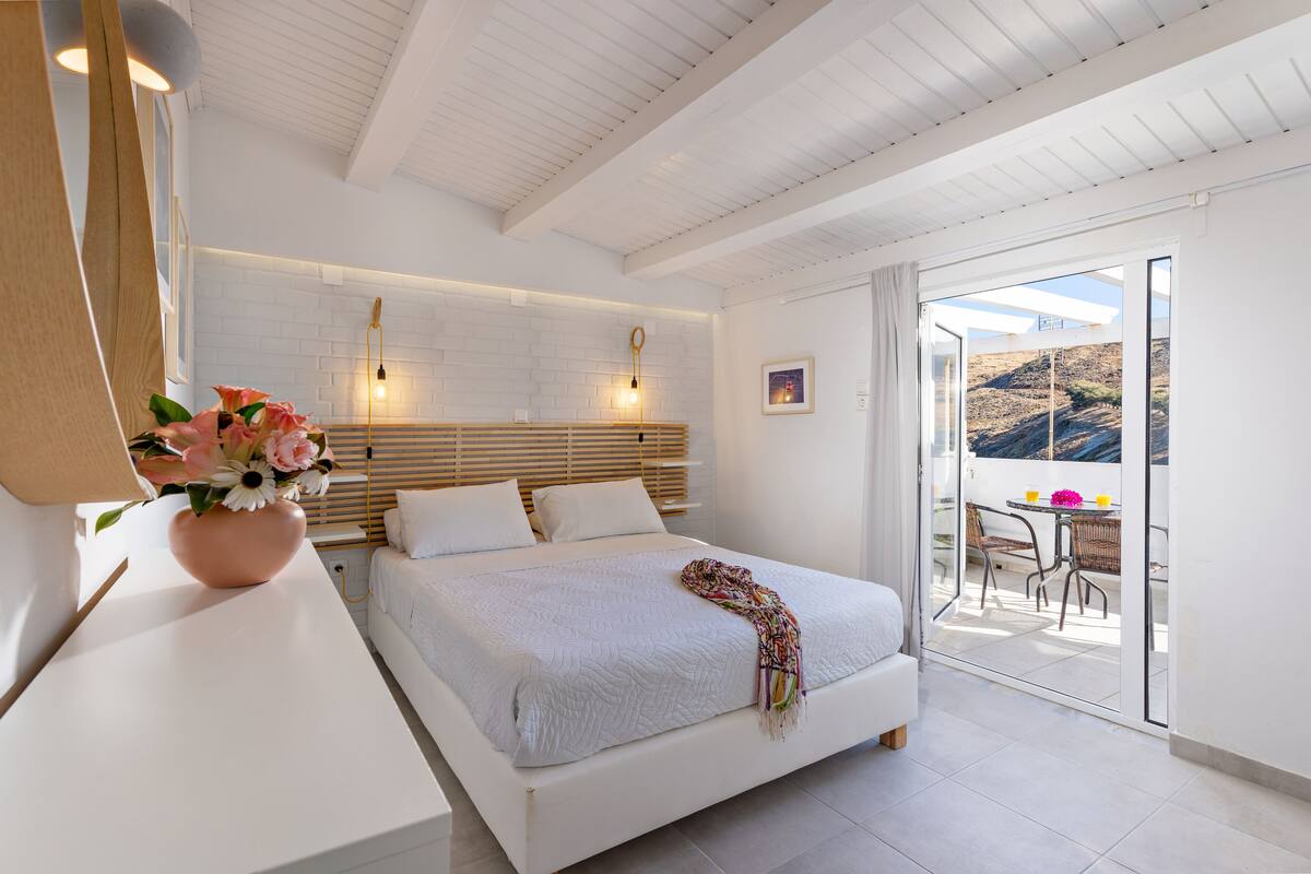A modern bedroom with a large bed, a bedside table with a vase of flowers, a white headboard, and a balcony with a table and chairs.