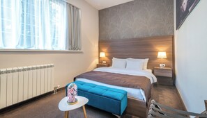 A hotel room with a bed, two bedside lamps, a radiator, a window with curtains, a blue bench, and a small table with a decorative item. - Veneti Suites (Belgrade)