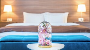 A bed with a blue upholstered headboard, a white pillow, and a glass dome containing pink flowers. There are two bedside lamps on either side of the bed.