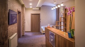 A hotel reception area with a wooden reception desk, a TV screen, a wall-mounted air conditioner, and a decorative vase with flowers.