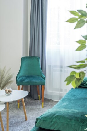 A room with a teal upholstered sofa, a small round table with a glass vase, and a chair with wooden legs.
