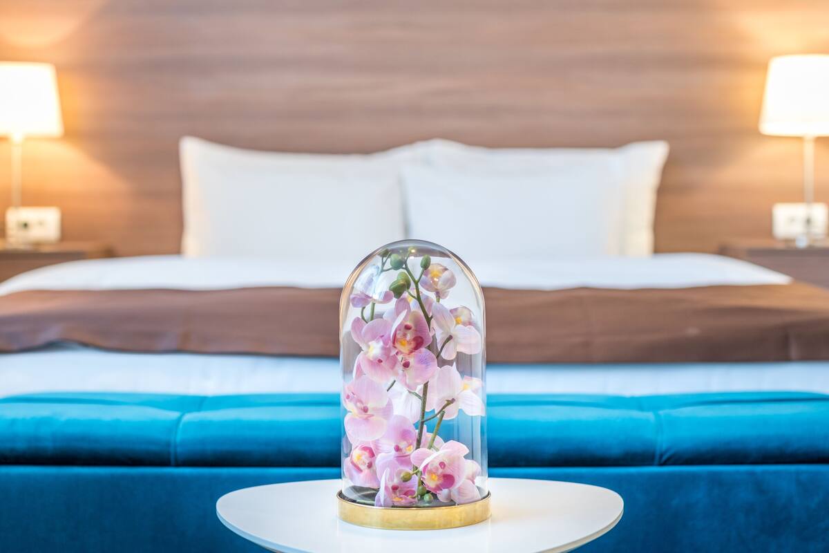 A bed with white linens, a blue upholstered bench, and a clear dome displaying pink orchids.