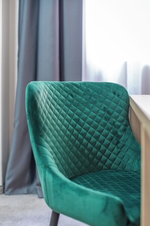 A green upholstered chair with a quilted pattern, positioned in a room with a window and curtains. - Veneti Suites (Belgrade)