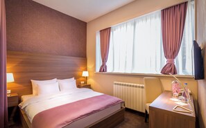Ett hotellrum med en säng, sänglampor, ett skrivbord med en stol, ett element och ett fönster med gardiner.