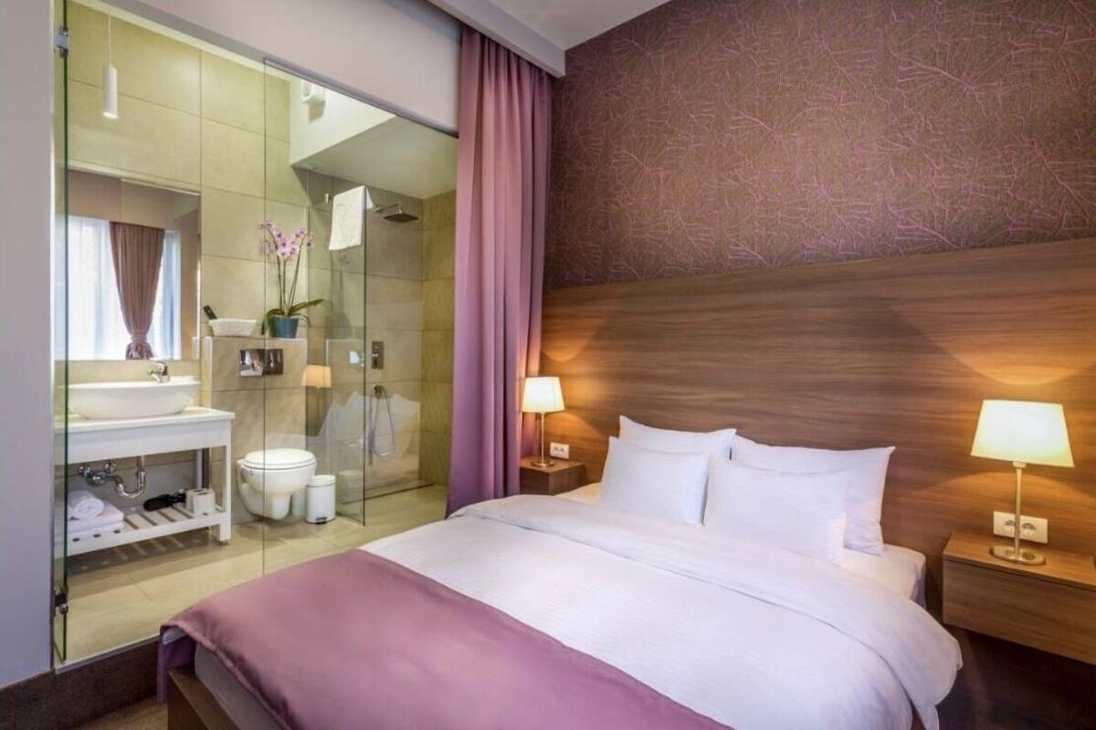 A hotel room with a bed, bedside lamps, a nightstand, a glass shower enclosure, a vanity with a sink, and a mirror.