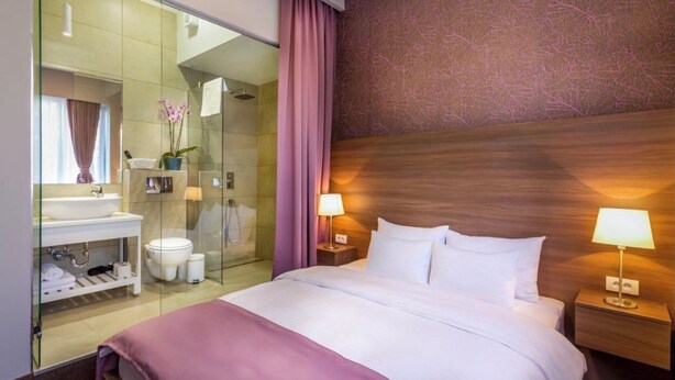 A hotel room with a bed, bedside lamps, a nightstand, a glass shower enclosure, a vanity with a sink, and a mirror.