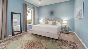 Une chambre à coucher avec un grand lit, un ventilateur de plafond, une fenêtre avec des rideaux, un tapis et une table de chevet avec une lampe.