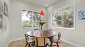 Une salle à manger avec une table ronde en bois, six chaises, un luminaire suspendu rouge et de grandes fenêtres donnant sur la forêt.