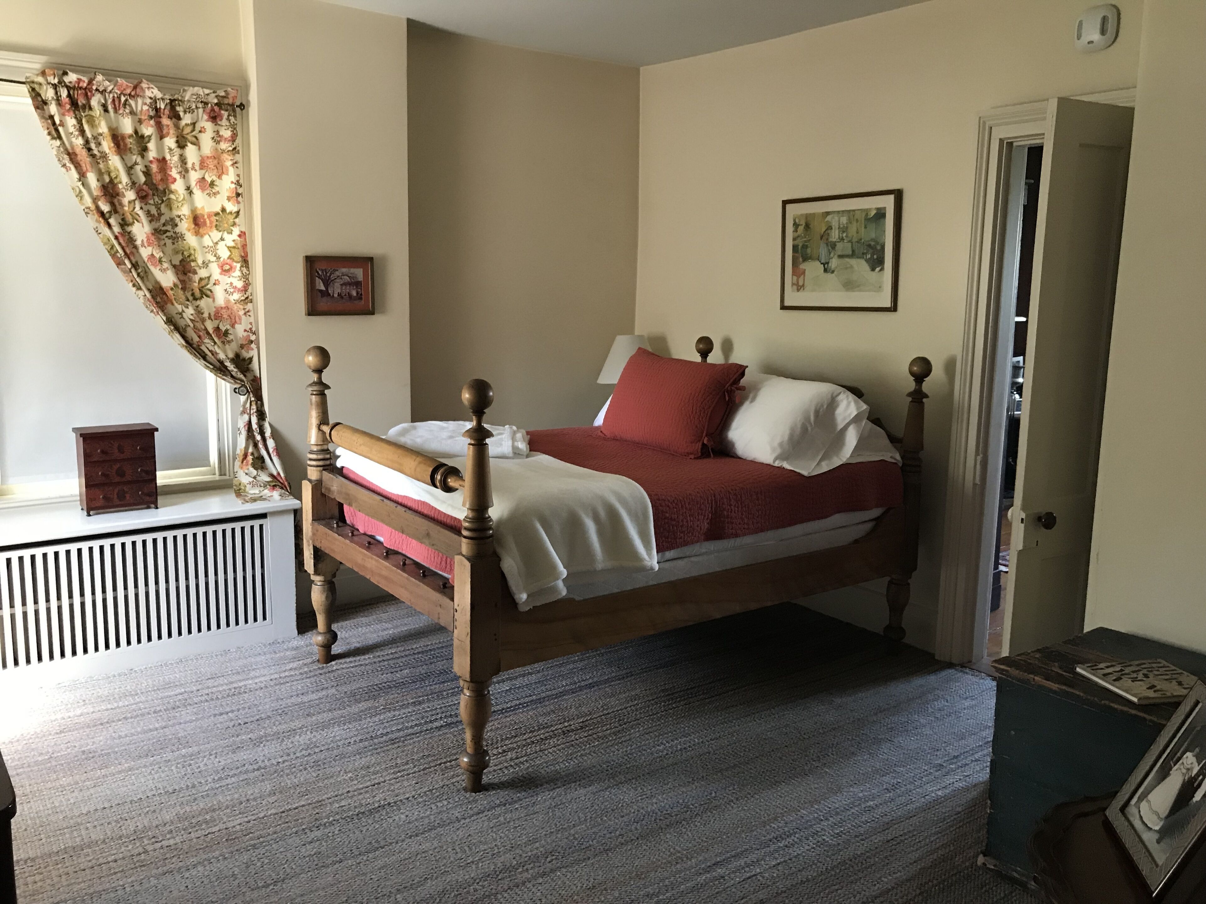 A bedroom with a wooden bed, a window with floral curtains, a radiator, and a door leading to another room.