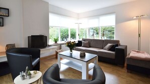 Een moderne woonkamer met een bank, fauteuils, een salontafel en een televisie. Grote ramen bieden uitzicht op groen buiten.