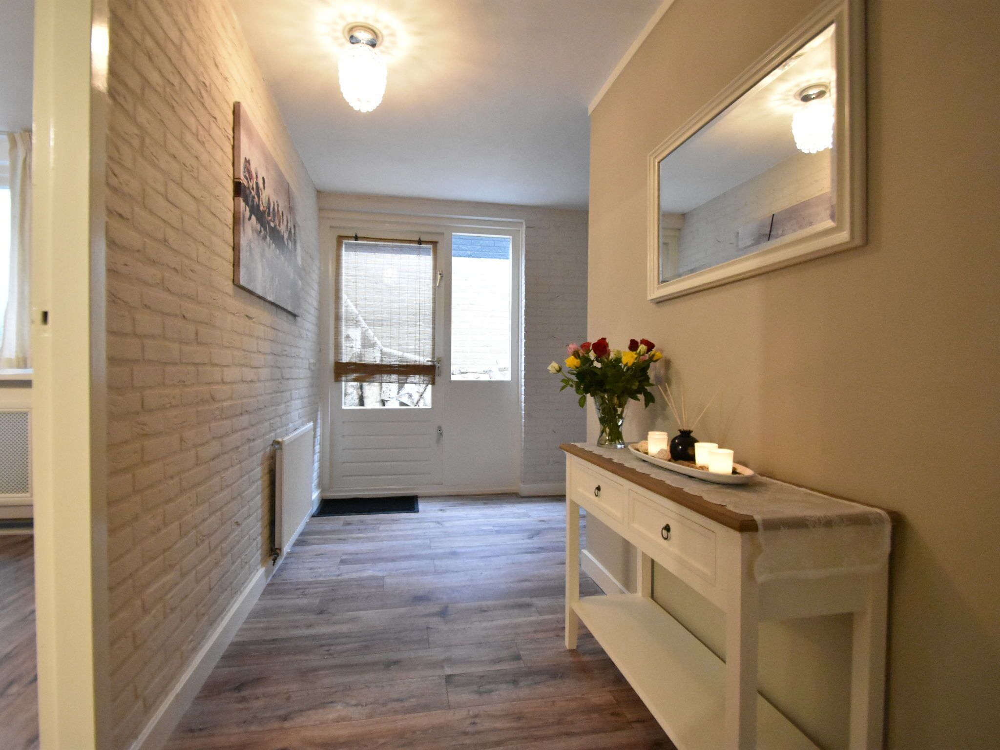 A hallway with a wooden floor, a white door, a mirror on the wall, and a vase with flowers on a console table.