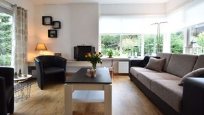 Een moderne woonkamer met een bank, een salontafel, een televisie en een uitzicht op groen door grote ramen.
