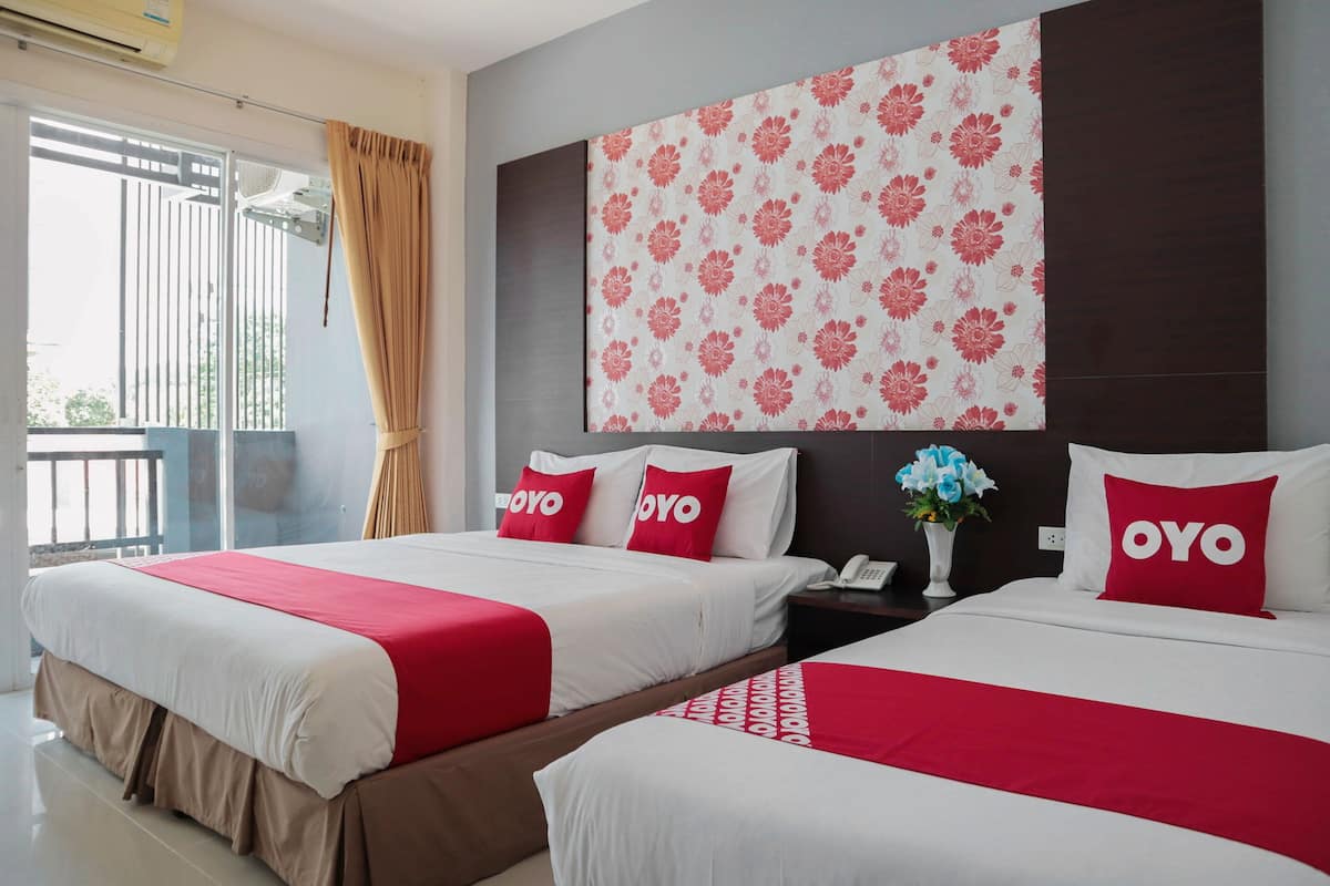 Habitación de hotel con dos camas, cada una con una colcha roja y blanca y almohadas a juego. La habitación tiene un ventanal con cortinas, un aire acondicionado de pared y un papel tapiz decorativo con motivos florales.
