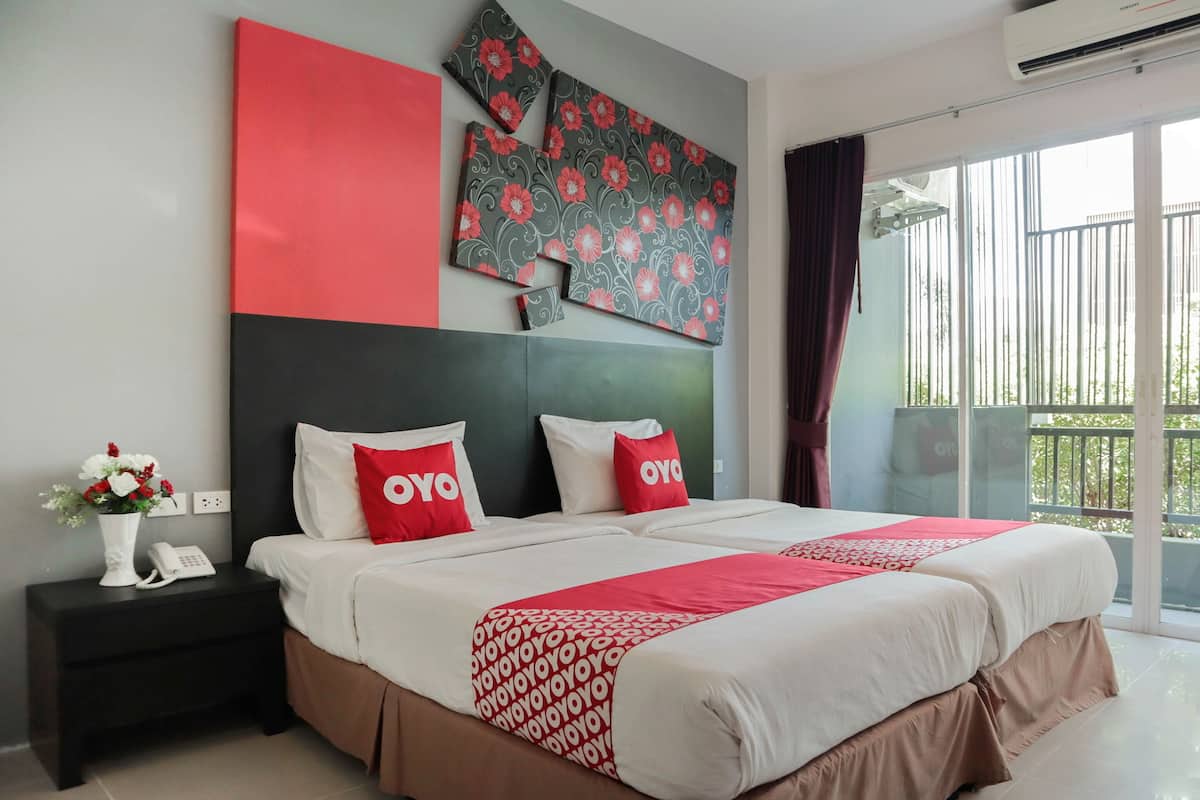 Habitación de hotel con una cama grande, dos almohadas rojas con la marca 'OYO', una mesita de noche negra con un jarrón blanco y un teléfono, y un balcón con vistas.