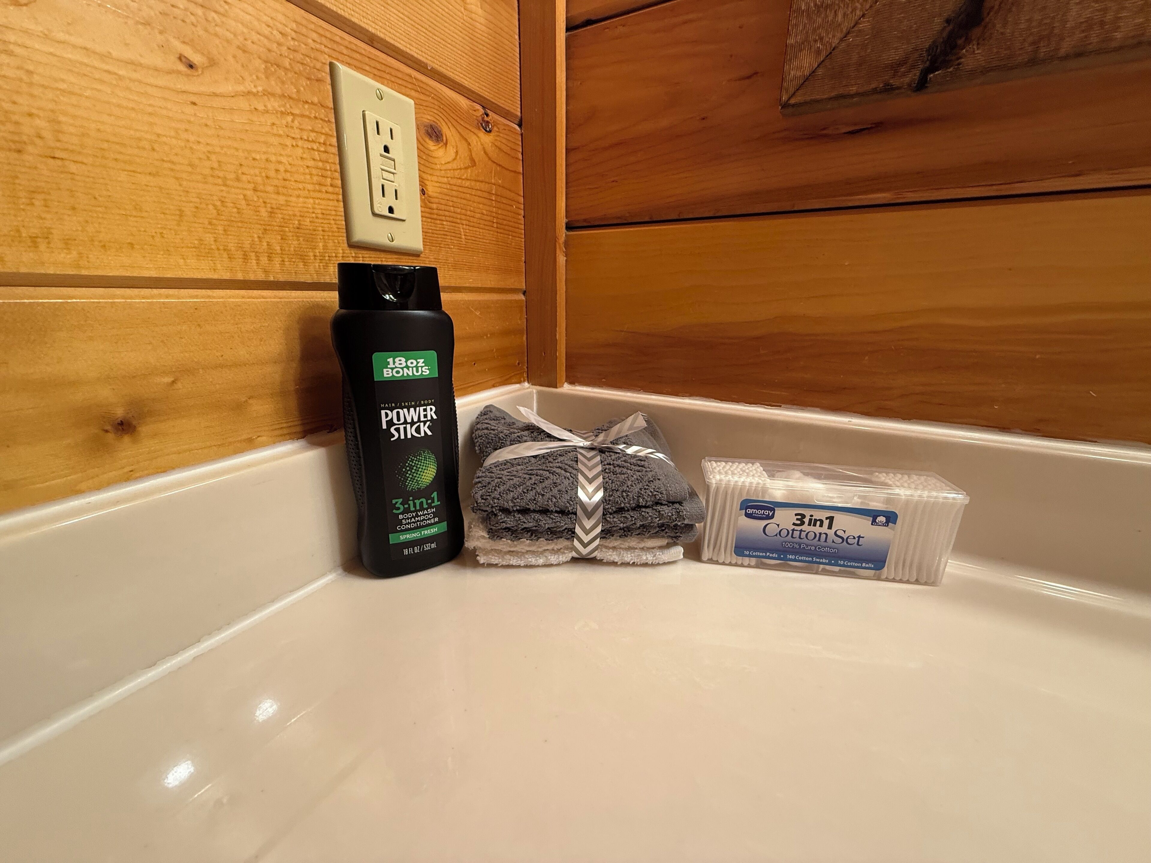 A bathroom with a wooden wall, a white sink, a bottle of Power Stick shampoo, a stack of gray towels, and a set of cotton pads.