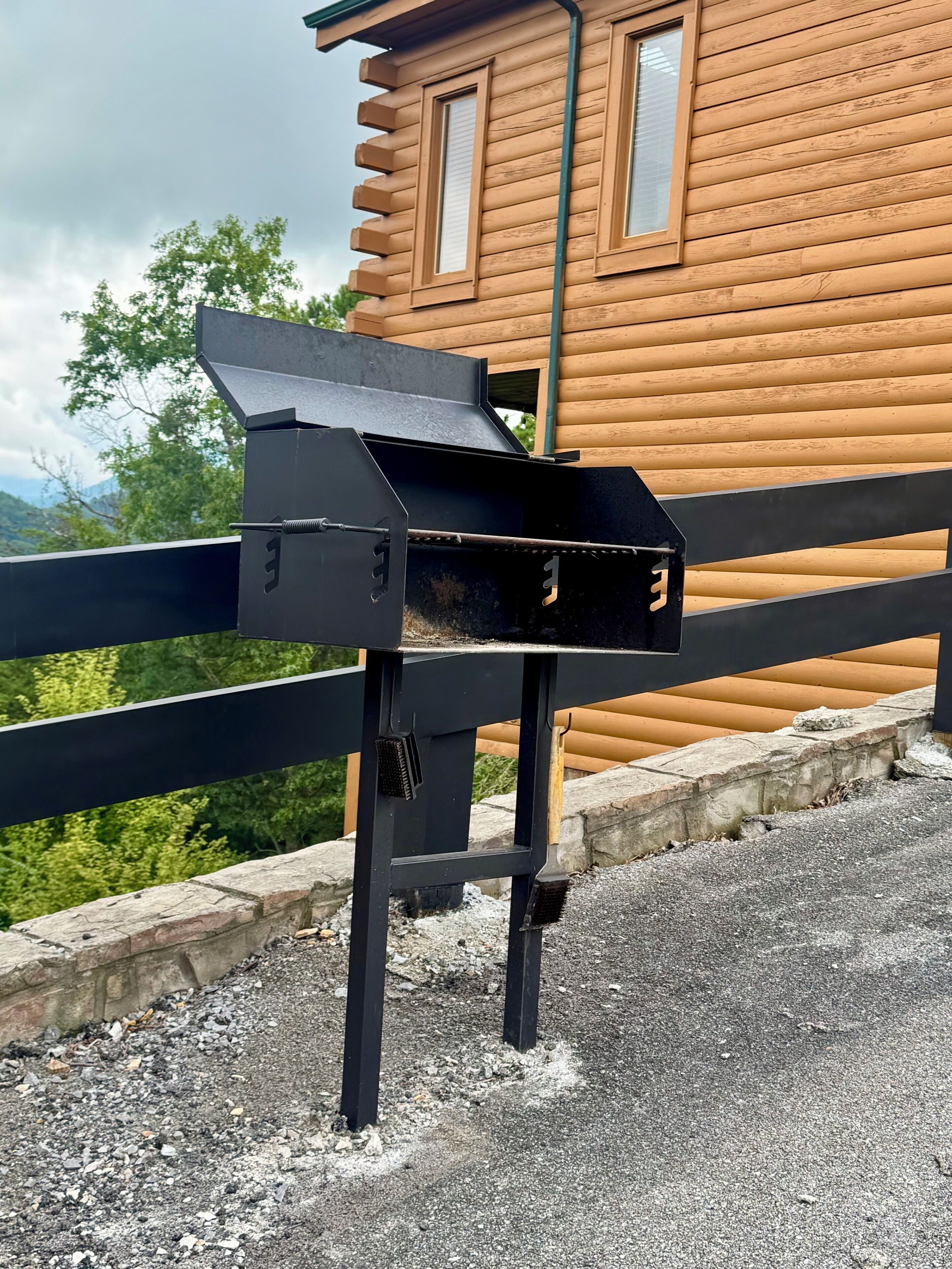 A black grill attached to a metal stand, positioned on a paved surface with a wooden building and a black fence in the background.