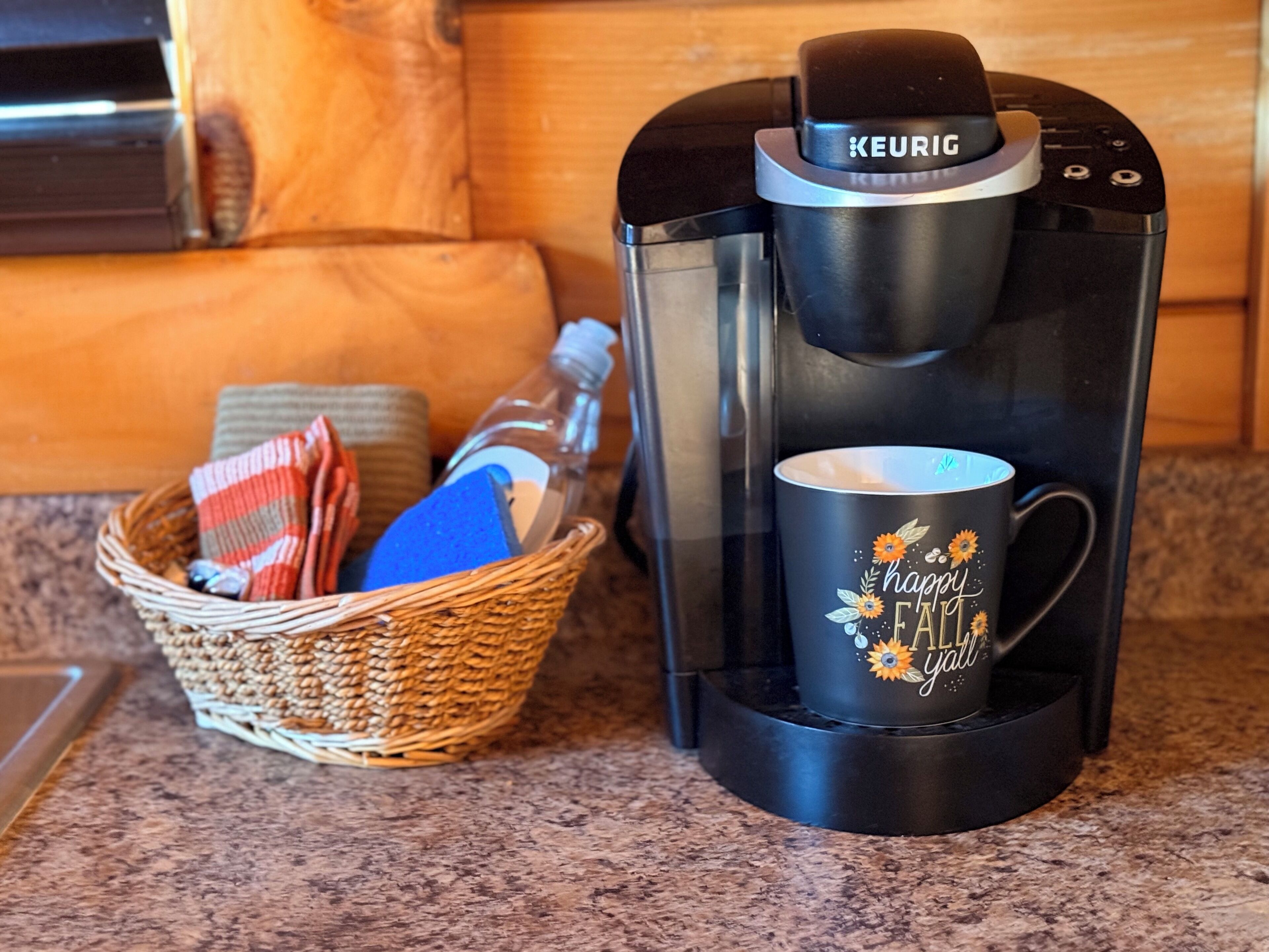 En Keurig-kaffemaskine med en kop på drypbakken, en fletkurv med et rødt og hvidt ternet stof, og et blåt objekt.