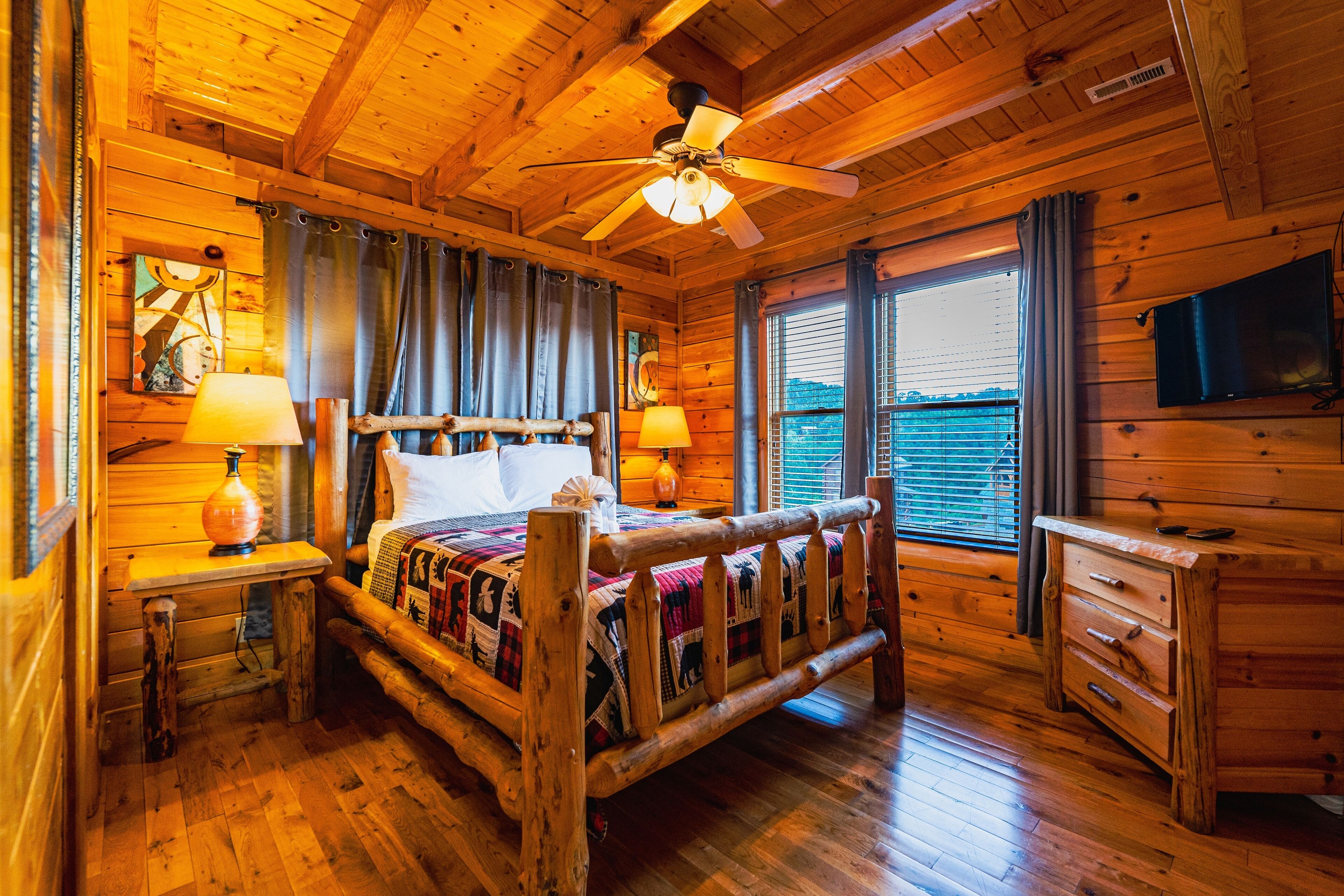 Un dormitorio de cabaña de madera con cama, mesitas de noche, cómoda, ventilador de techo y ventana con cortinas.
