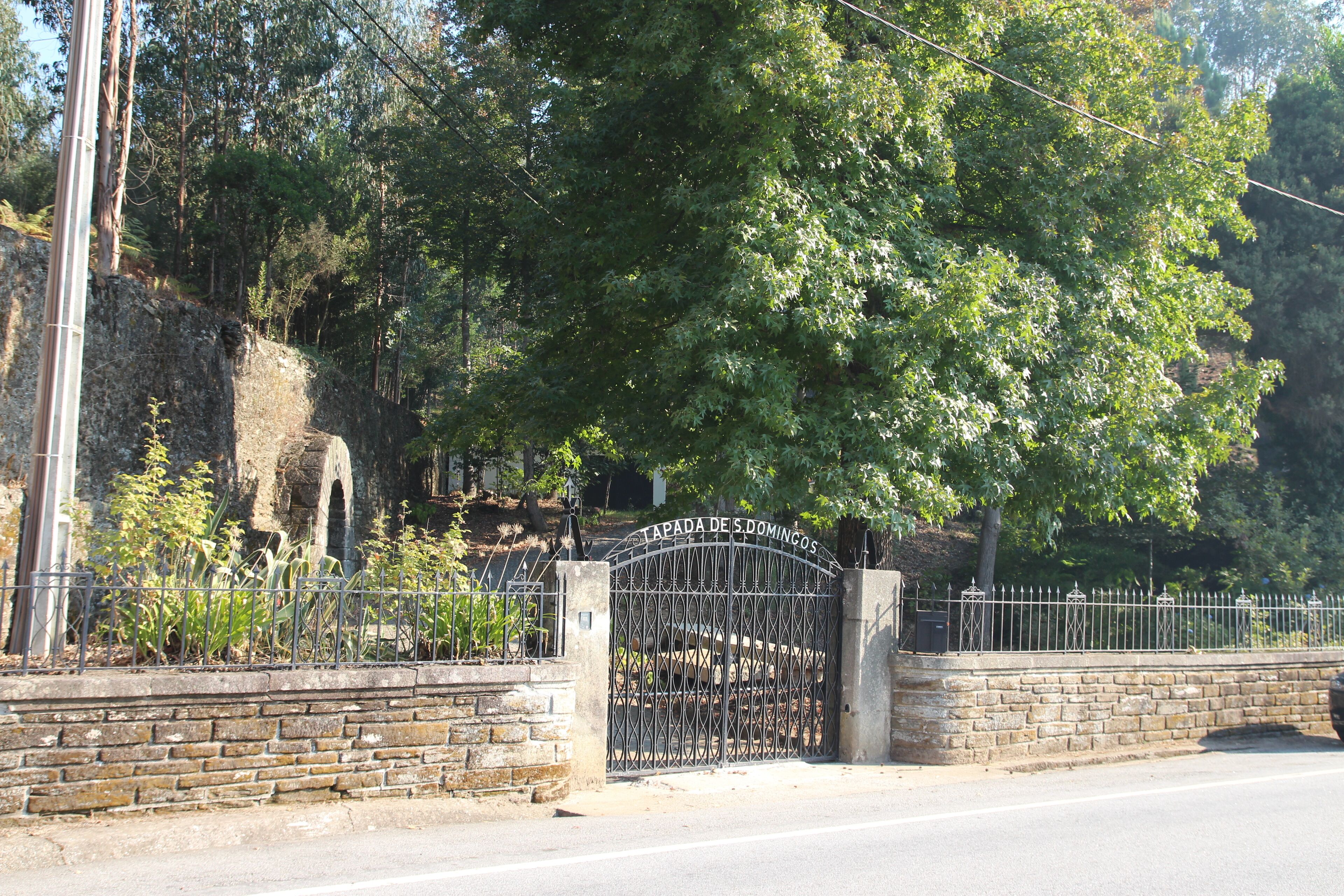 Un mur de pierre surmonté d’un portail en métal portant l’inscription « APADA DE S. DOMINGOS » et un escalier en pierre menant à une zone rocheuse bordée d’arbres.