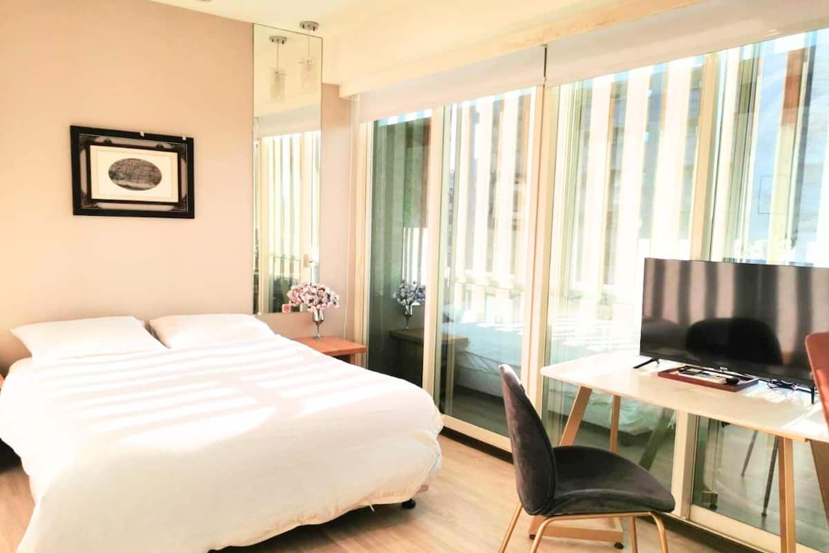 A hotel room with a bed, a desk with a chair, a television, and a large window with curtains.