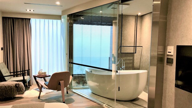 Une chambre d’hôtel moderne dotée d’un grand lit, d’un coin salon avec un fauteuil et une table, d’une grande baignoire et d’une paroi en verre.