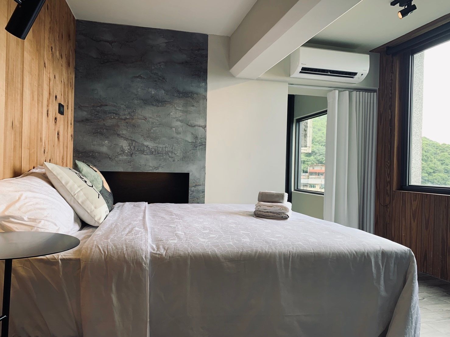 Quarto moderno com cama grande, cabeceira de madeira, parede de concreto como destaque e vista para o verde lá fora.