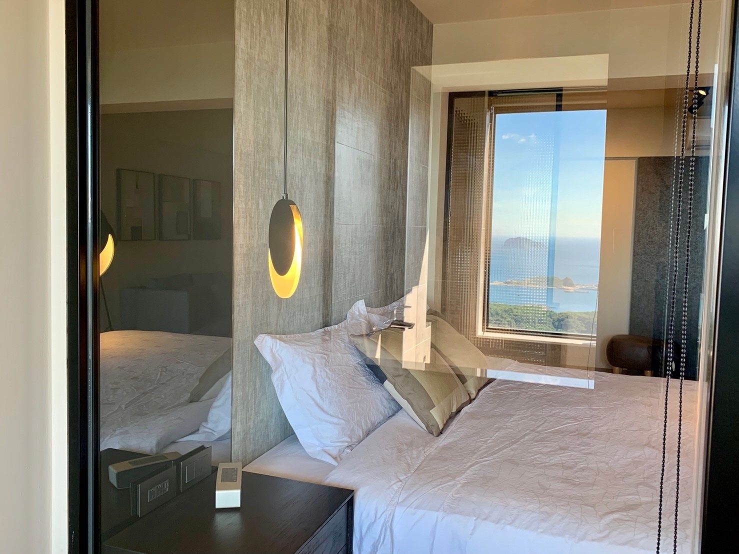 Un dormitorio moderno con una cama grande, una mesita de noche, una lámpara colgante y una ventana con vistas al mar y al paisaje urbano.