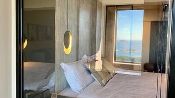 Un dormitorio moderno con una cama grande, una mesita de noche, una lámpara colgante y una ventana con vistas al mar y al paisaje urbano.