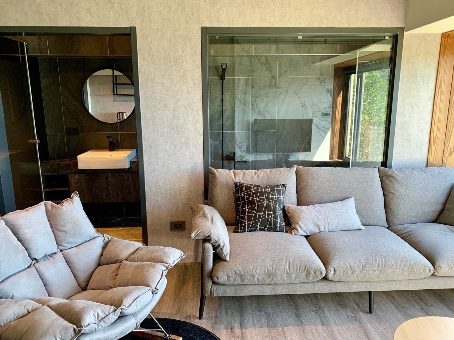 A modern living room with a sofa and armchair, a round mirror, and a glass shower enclosure.