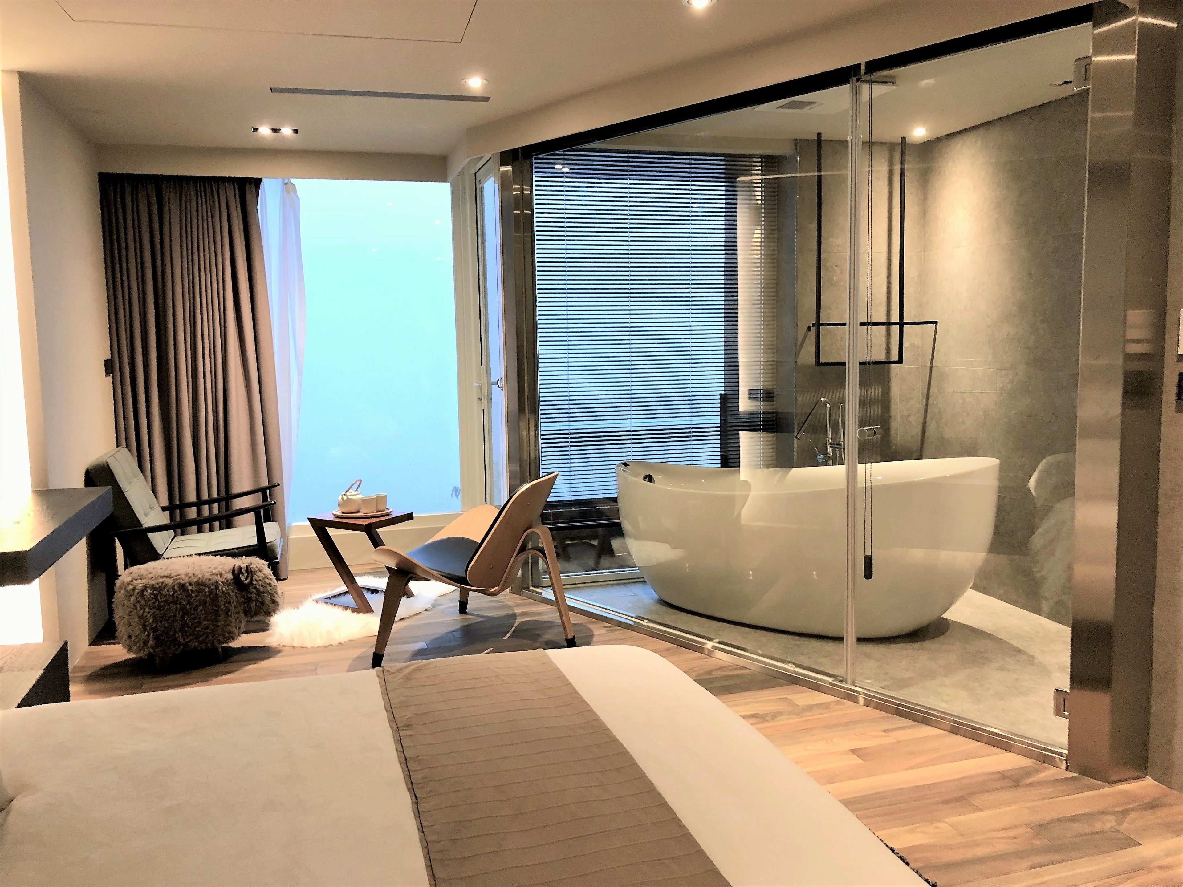 A modern bedroom with a large bed, a glass-enclosed bathtub, and a seating area with a chair and table.