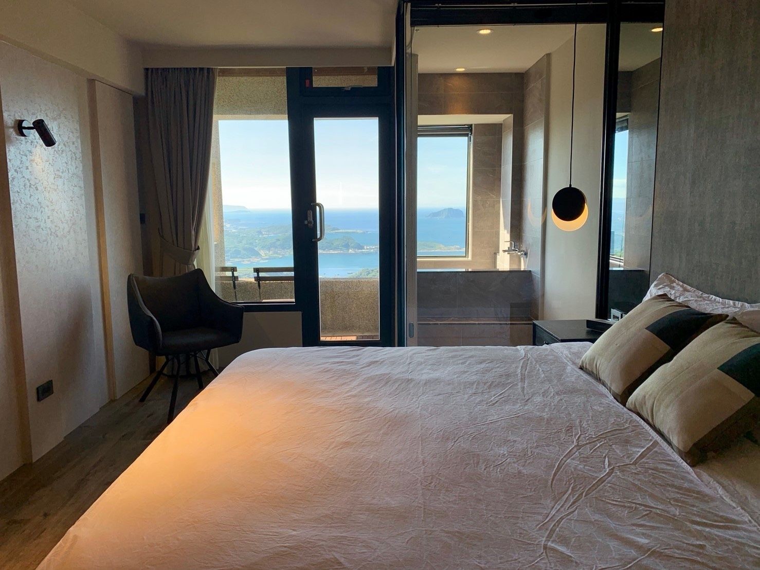 A hotel room with a large bed, a chair, a bathroom with a view, and a balcony.