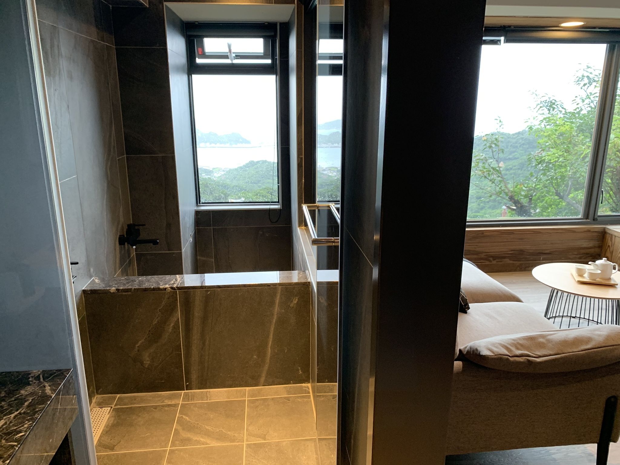 A modern bathroom with a shower, a bathtub, and a window offering a view of greenery.