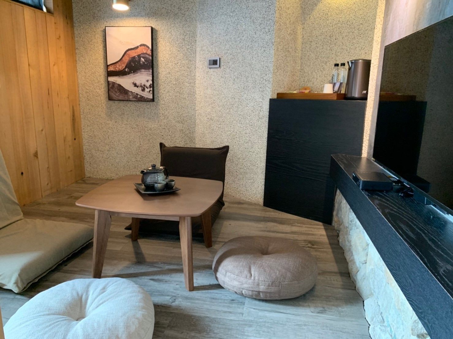 A cozy room with a wooden table, a cushioned bench, a small round cushion, a painting on the wall, and a TV on a black stand.