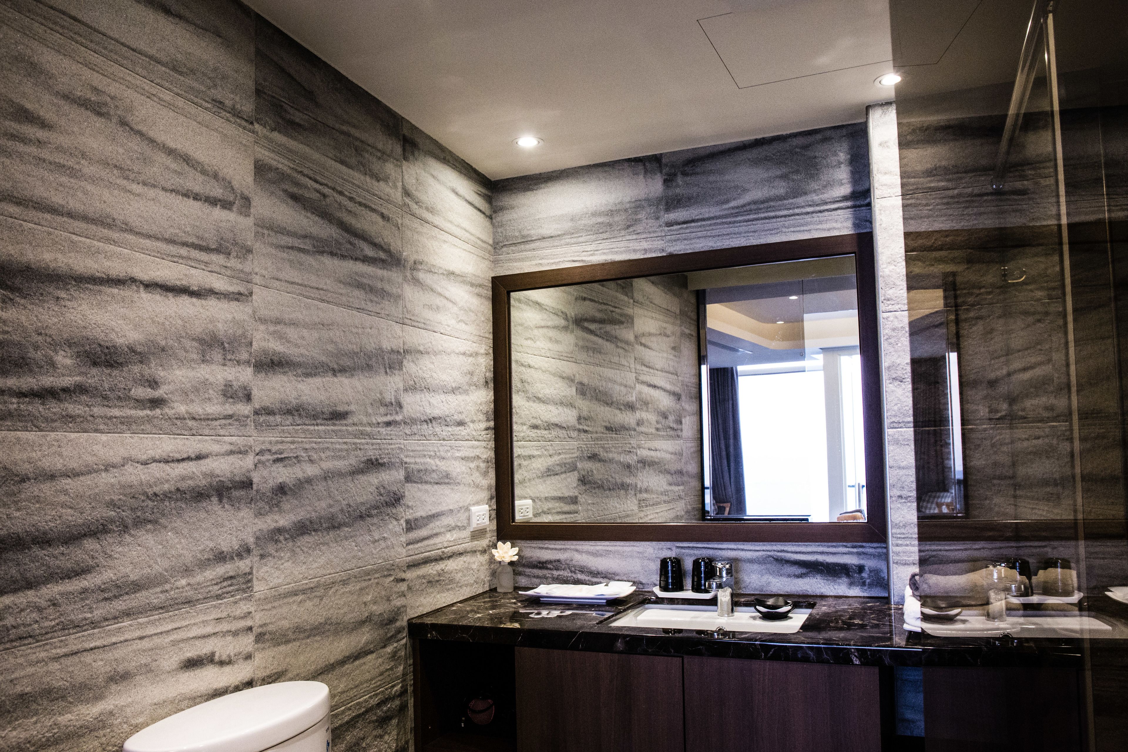 A modern bathroom with a marble wall, a large mirror, and a dark countertop.