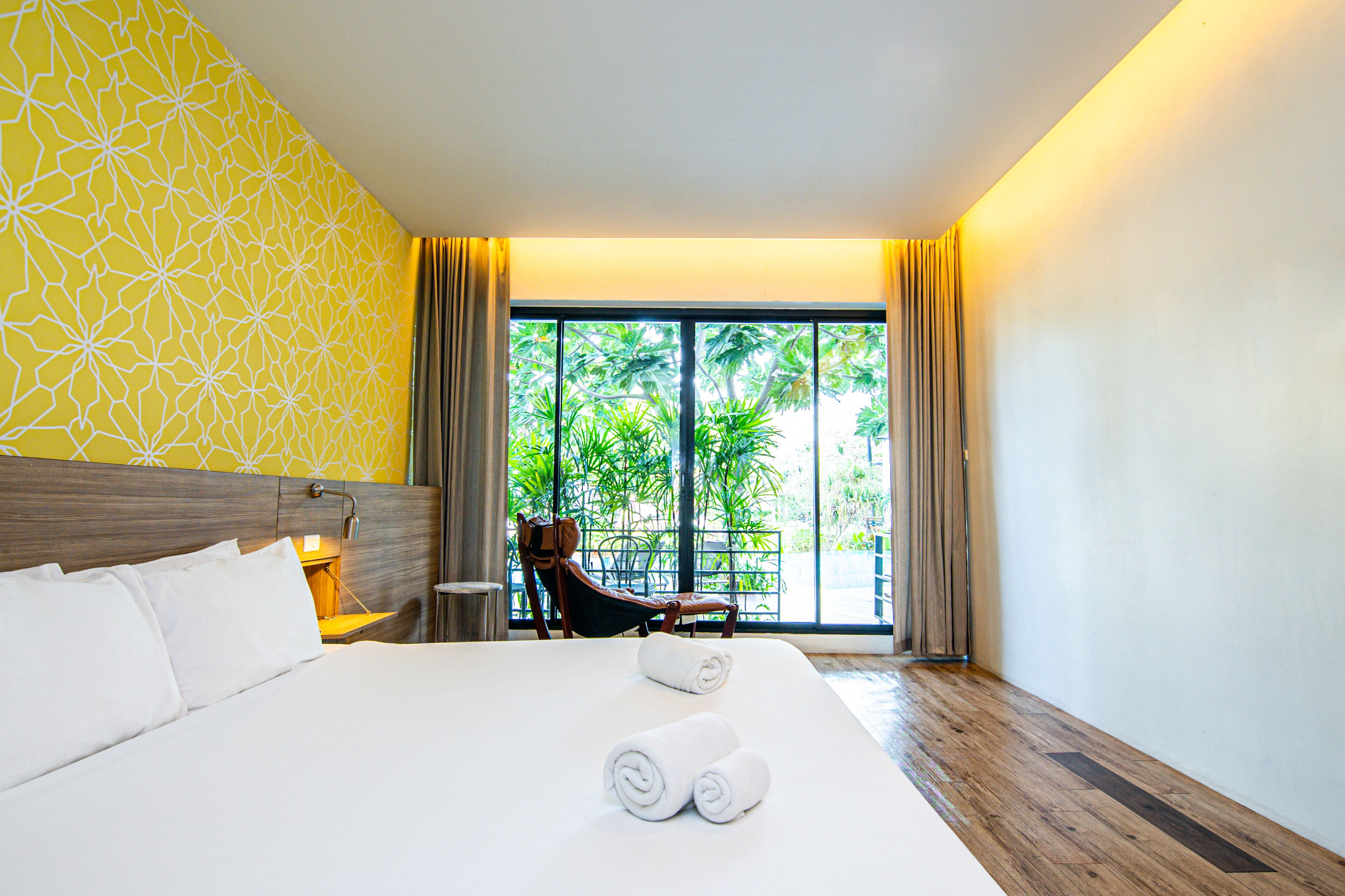 A hotel room with a large bed, a wooden headboard, a chair, and a view of greenery through the window.